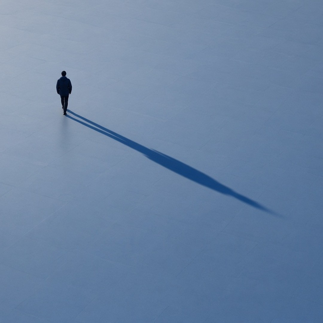 From high above, one small walker crosses an empty blue-grey plaza, trailed by a shadow many times their size